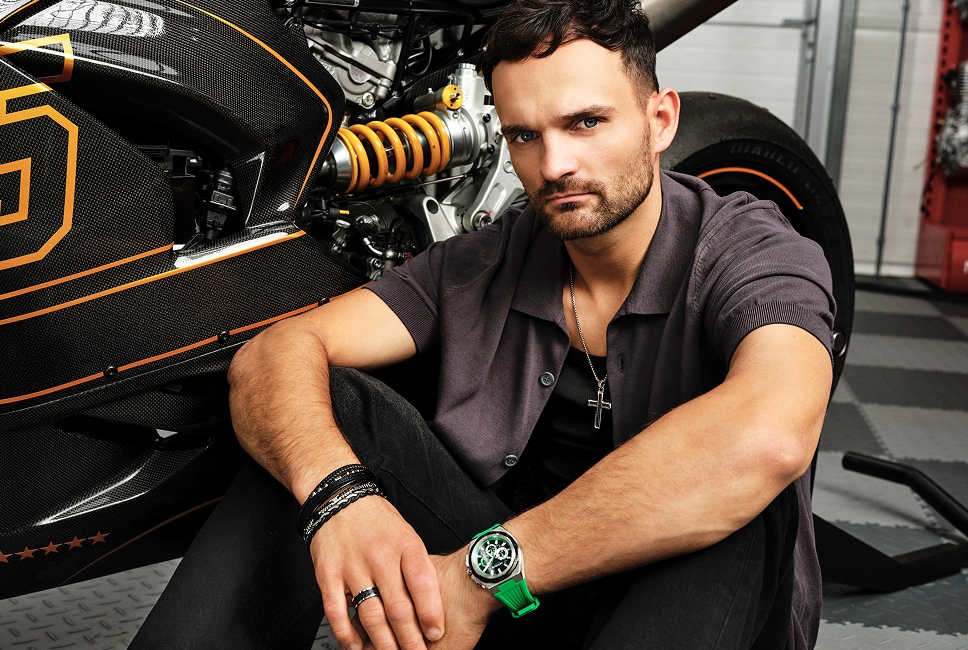 Bartosz Zmarzlik předvádí šperky a hodinky značky Aztorin v pozadí je motocykl