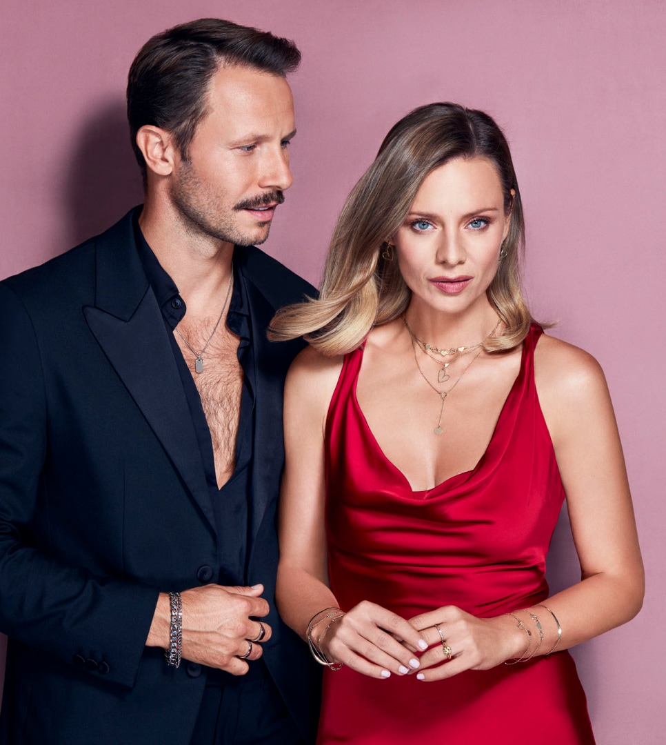 Magdalena Boczarska a Mateusz Banasiuk představují stříbrné dámské a pánské šperky na růžovém pozadí - náušnice, náhrdelníky, prsteny, náramky