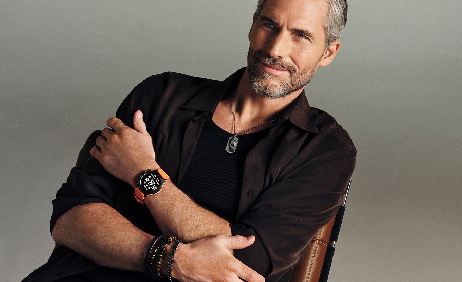 A man leaning against a chair, dressed in black, shows a set of jewelry and a men's watch.