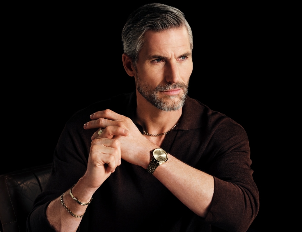 Mężczyzna w brązowym swetrze prezentujący złoty zegarek oraz złotą biżuterię