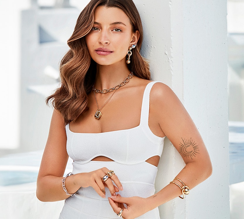 Julia Wieniawa ubrana na biało na tle budynków prezentująca srebrną oraz pozłacaną biżuterię z kolekcji Światłocienie by Julia Wieniawa