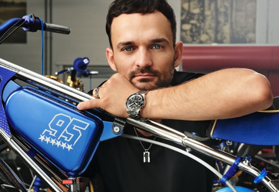 Mężczyzna opierający się o niebieski motocykl żużlowy z numerem 95, prezentujący zegarek i naszyjnik