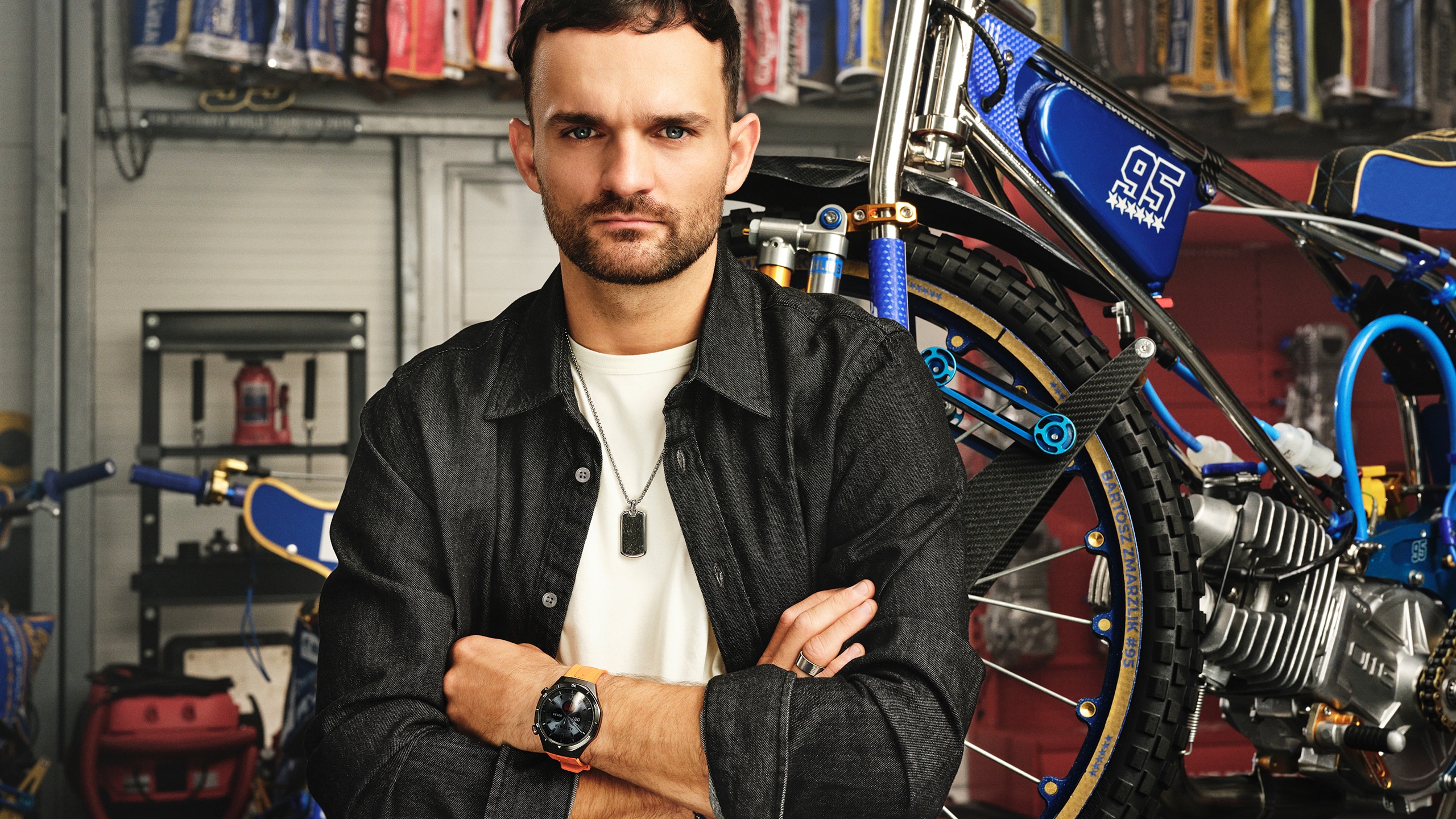 Mężczyzna w czarnej koszuli stojący obok motocykla żużlowego, prezentujący smartwatch Aztorin na pomarańczowym pasku