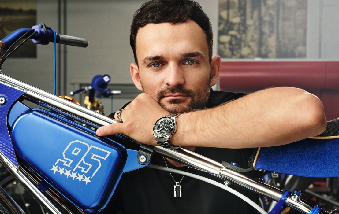 Bartosz Zmarzlik z luksusowym zegarkiem Rado HyperChrome Chronograph, opierający się o żużlowy motocykl