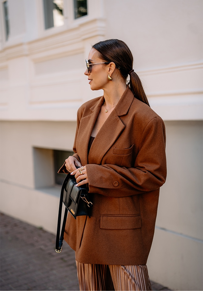 Kobieta w brązowej marynarce prezentująca złote kolczyki, naczyjnik oraz pierścionki