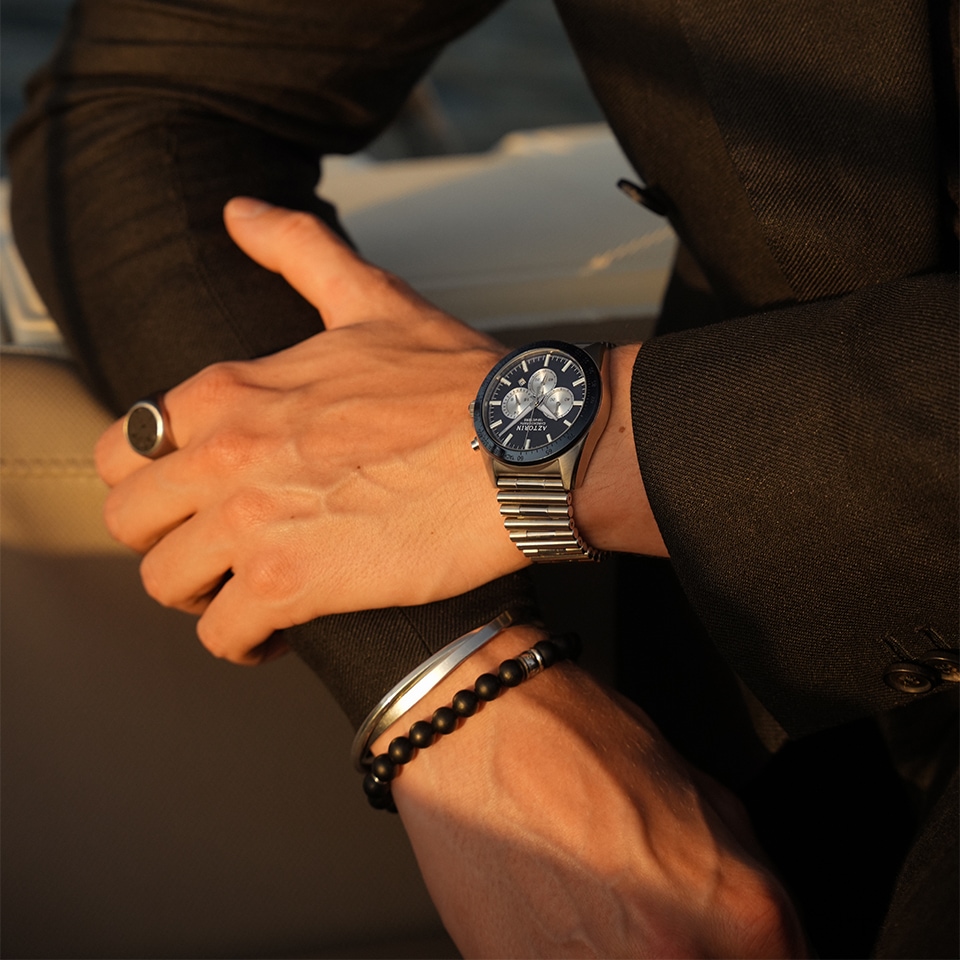 Mężczyzna ubrany w czarną koszulę prezentuje na tle zachodzącego słońca zegarek oraz biżuterię męską
