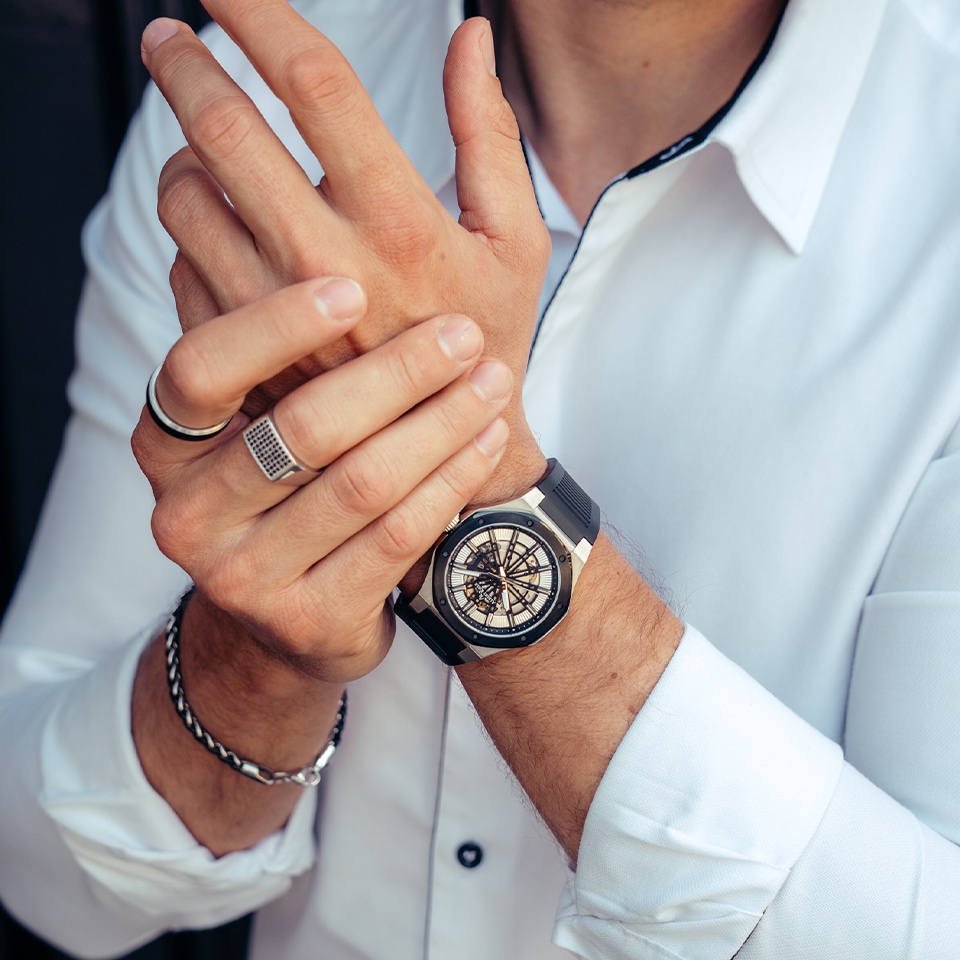 Mężczyzna ubrany w białą koszulę prezentujący zegarek oraz bransoletki i sygnety dobrane do eleganckiego stylu