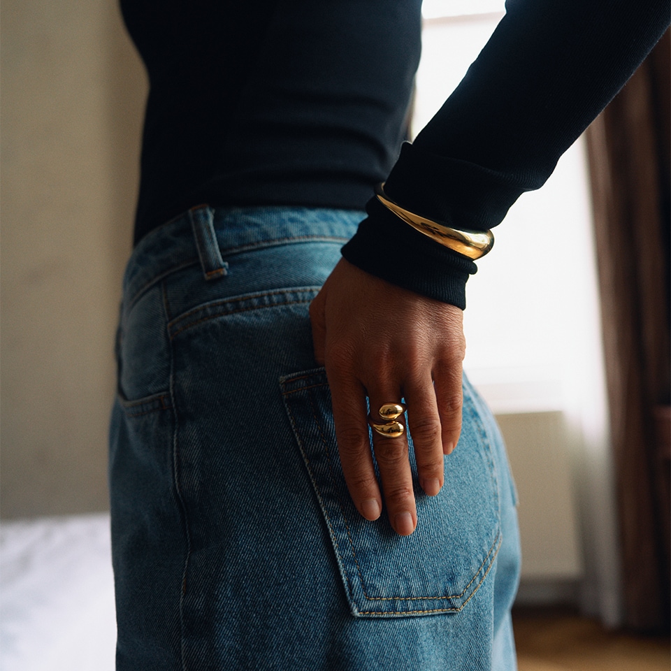 Kobieta ubrana w granatowy golf oraz niebieskie jeansy prezentuje złoty peirścionek oraz bransoletkę
