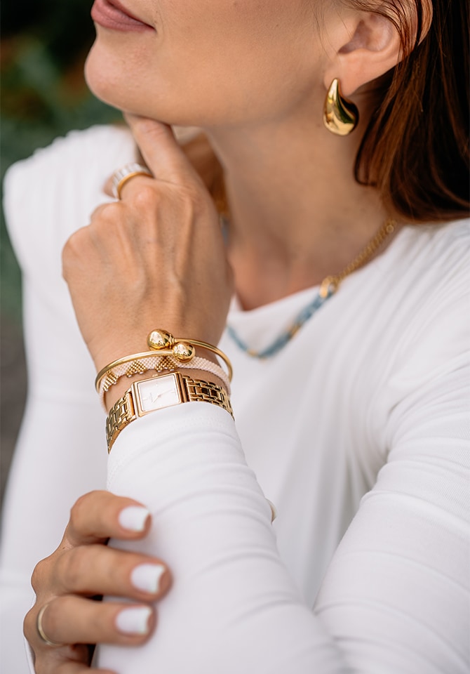 kobieta ubrana w białą koszule prezentuje komplet biżuterii złotej wraz z zegarkiem 