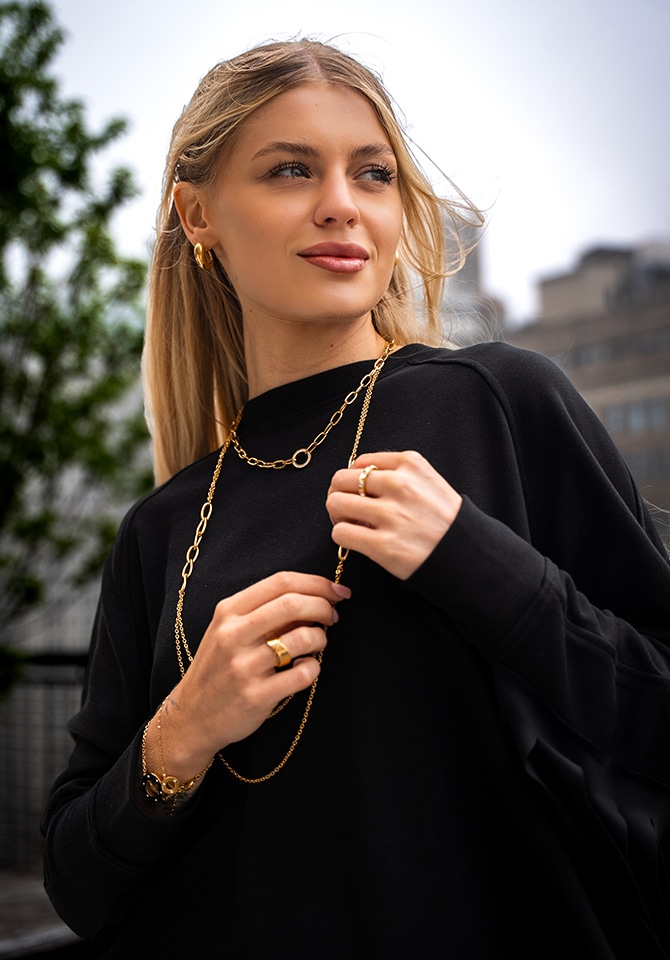 kobieta w długich blond włosach i czarnym dresowym komplecie prezentuje złota biżuterię - naszyjnik, kolczyki, pierścionki i bransoletki