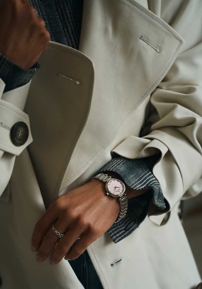 Kobieta w beżowym długim płaszczu jesiennym prezentyje zegarek z bransoletą ze stali szlachetnej