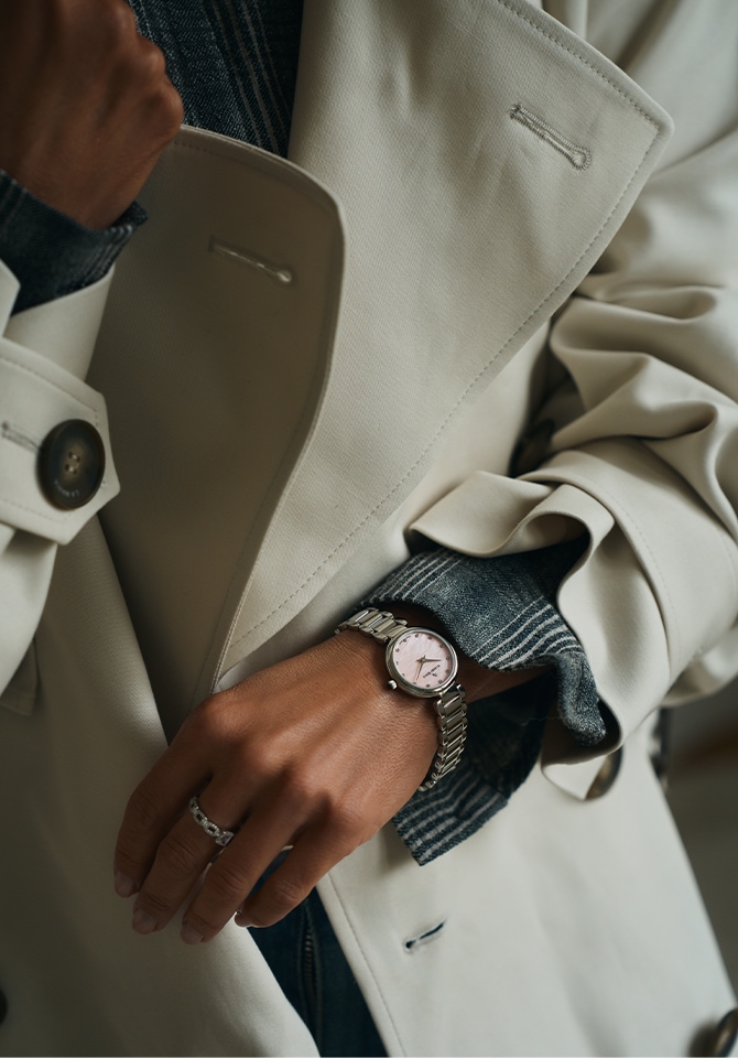 Kobieta w beżowym długim płaszczu jesiennym prezentyje zegarek z bransoletą ze stali szlachetnej