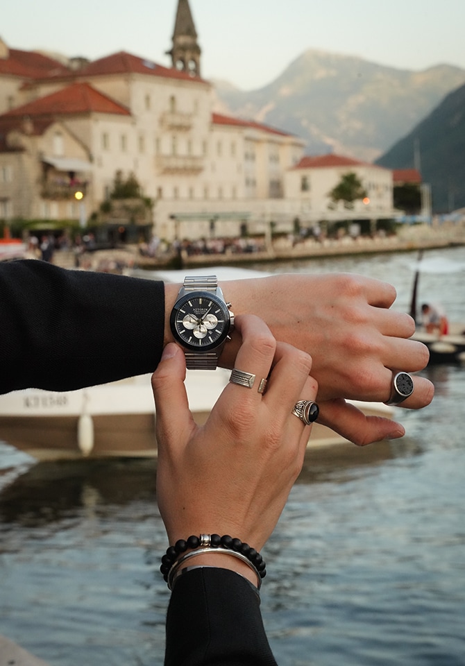 Mężczyzna prezntujący zegarek, bransoletki oraz sygnety męskie - w tle góry, budynki i chaos uliczny