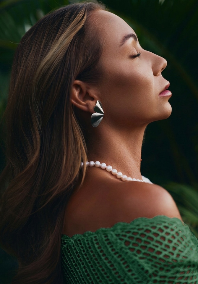 Kobieta w zielonej sukeince prezentuje kolczyki srebrne o nieregularnych kształtach oraz naszyjnik z perłami