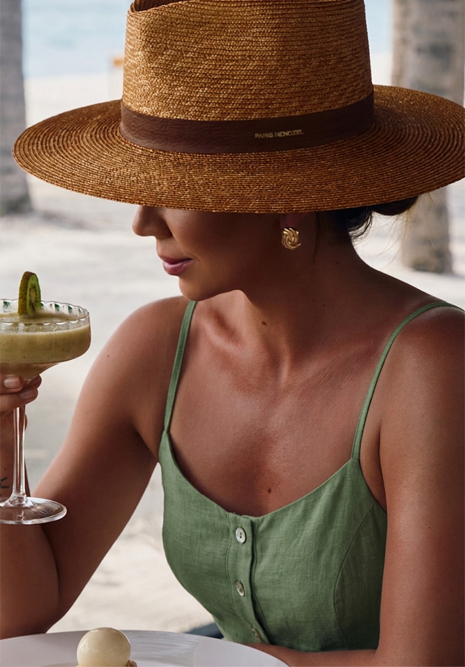 Kobieta w zielonej sukeince i słomianym kapeluszu prezentuje kolczyki pozłacane