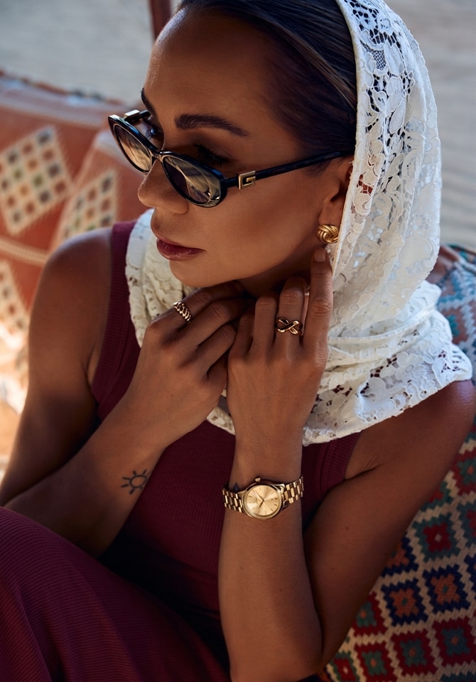 Modelka ubrana w bordową suknie przyozdobioną złotą biżuterią oraz zegarkiem i czarnymi okularami przeciwsłonecznymi