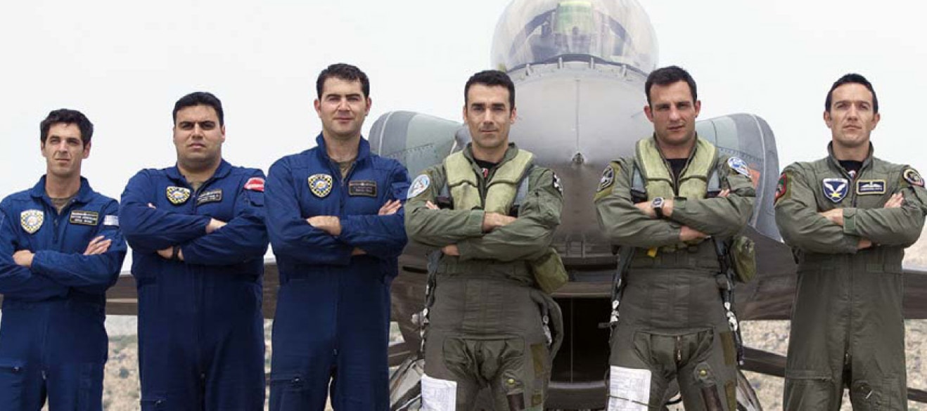 Grupa sześciu pilotów w kombinezonach lotniczych stoi przed wojskowym samolotem odrzutowym