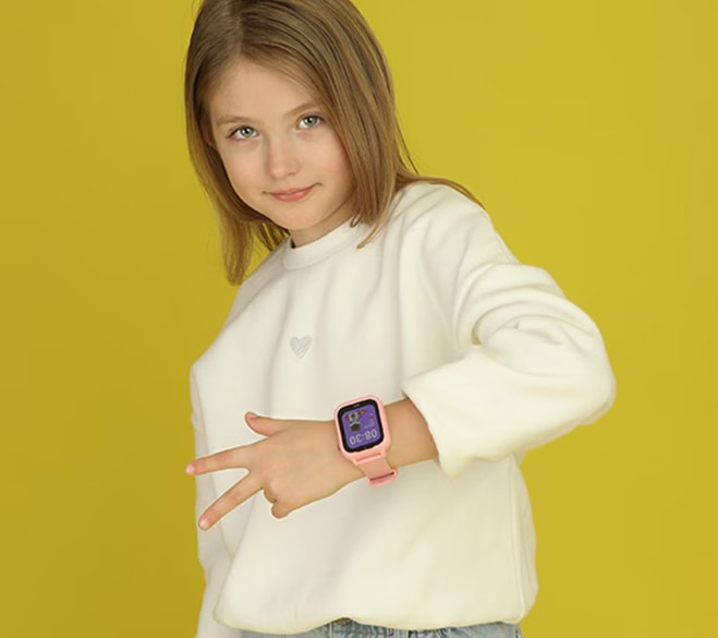 Dziewczynka o blond włosach stoi na tle żółtego tła i pokazuje różowy smartwatch na nadgarstku