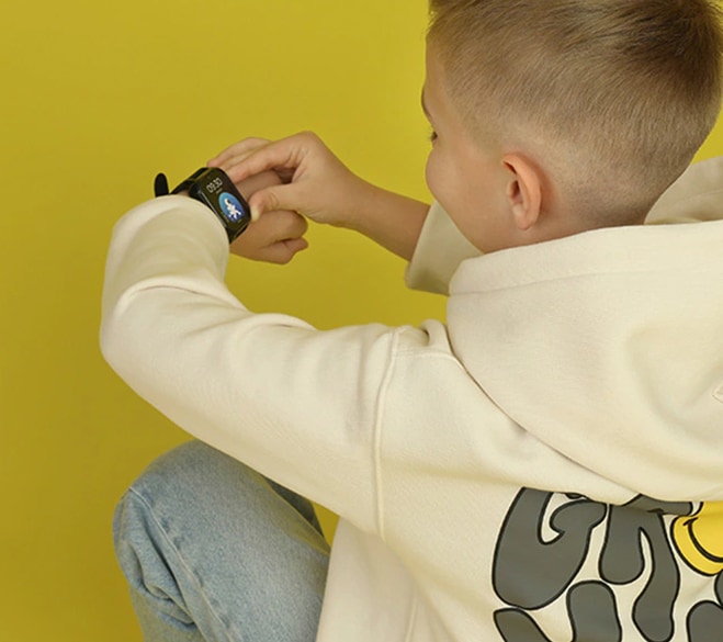 Chłopiec na żółtym tle ubrany w beżową bluze wpatrujący się w smartwatch założony na nadgarstku